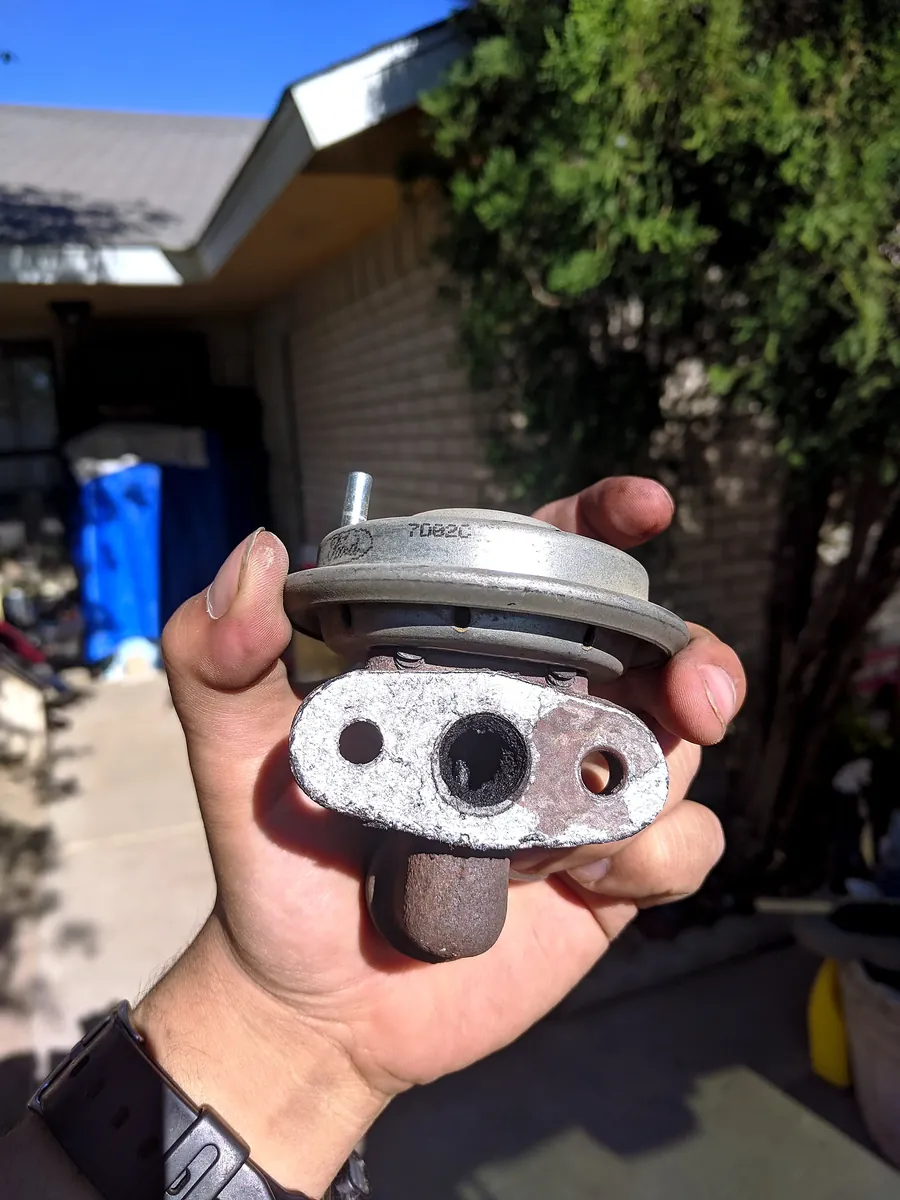 A Ford-stamped vacuum EGR valve held in a hand, mounting flange and carbon-clogged exhaust port facing the camera