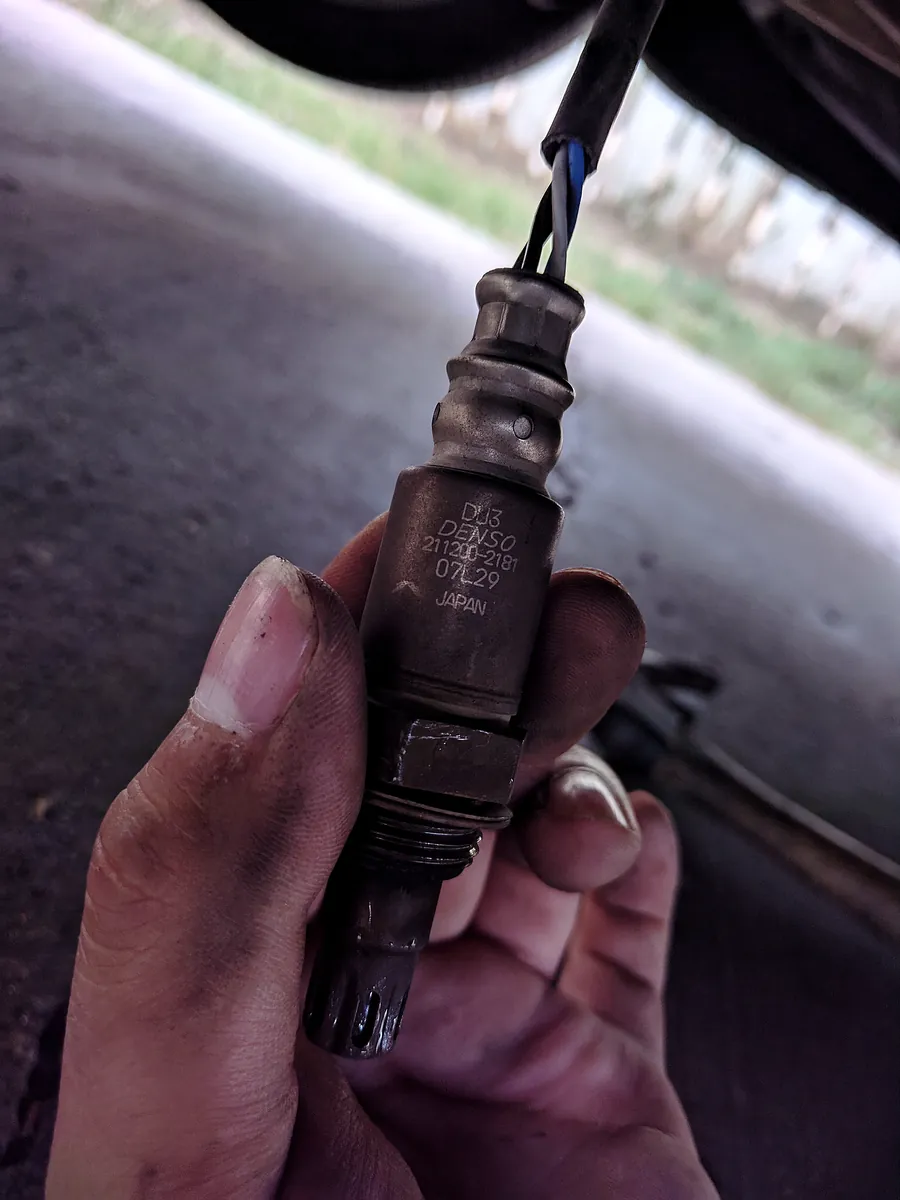 A used Denso oxygen sensor with its wiring, held in a hand under a car