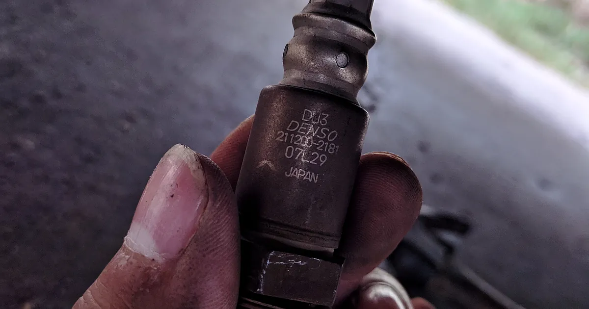 A used Denso oxygen sensor with its wiring, held in a hand under a car
