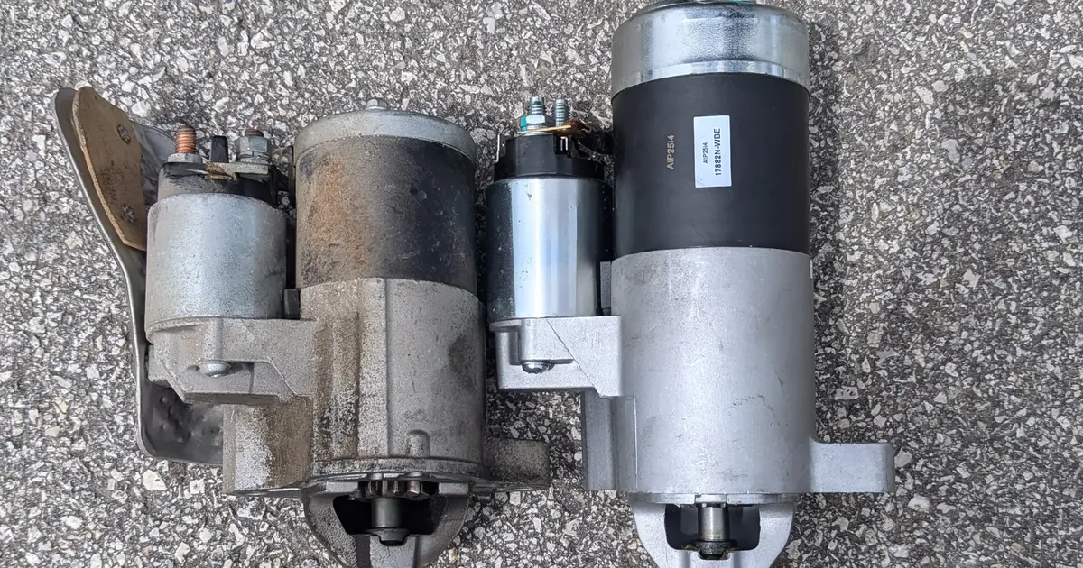 A worn, dirty starter motor lying next to a new starter on asphalt