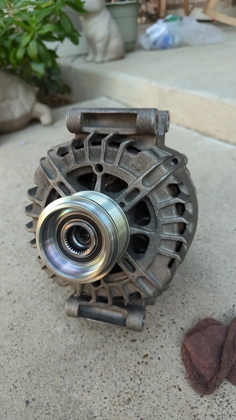 An alternator with its multi-groove pulley and clutch facing the camera, sitting on a concrete step